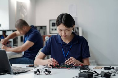 Modern teknoloji laboratuvarında elektronik bileşenler üzerinde yoğunlaşan yetenekli mühendislik lehimleme devresi. Arka planda başka bir iş yerinde çalışan meslektaşlar yer alıyor