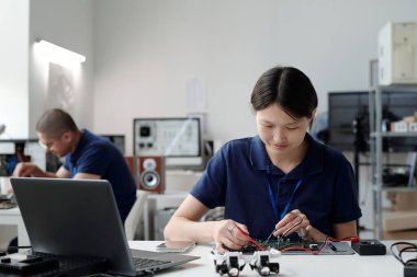 Modern laboratuarda otururken Asyalı mühendis devre kartını tamir ediyor. Çalışma geçmişi ve mevcut çeşitli elektronik ekipmanları içerir