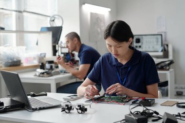 Modern ofis ortamında devre kartlarında çalışan mavi gömlekli mühendisler. Çeşitli elektronik aletler ve aletlerle çevrili karmaşık kablolama ve cihaz montajına odaklanan insanlar