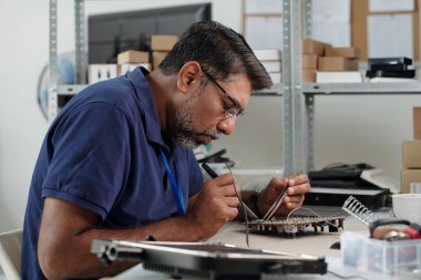 Çalışma alanındaki devre kartlarını tamir etmeye odaklanan bir adam. Kutular ve ekipmanlarla dolu raflar. Çalışma alanında görülebilen küçük elektronik bileşenleri kullanmada hassasiyet