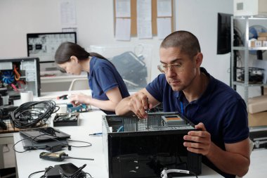 Kadın ve erkek teknoloji atölyesinde çalışıyor, bilgisayar donanımı ve elektronik cihazları tamir ediyor. Düzenli ortamda çeşitli araç ve gereçler kullanan odaklanmış profesyoneller