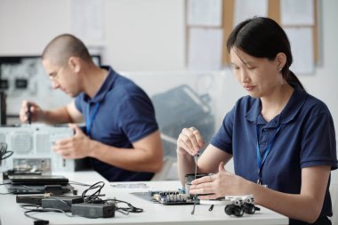 Teknoloji atölyesindeki tezgahta elektronik parçaları birleştiren iki teknisyen, karmaşık göreve odaklanarak, çalışmalarında hassasiyet ve uzmanlık gösteriyorlar.