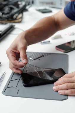 Teknoloji tezgahında özel aletler kullanarak kırılmış akıllı telefon ekranını tamir eden ellerin yakın çekimi. Birçok küçük bileşen ve elektronik cihaz etrafa saçılmış durumda.