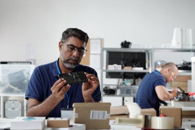 Teknoloji atölyesindeki mobil cihazı inceleyen mühendis çeşitli elektronik bileşenler ve cihazlarla dolu, ürünün kalitesini ve işlevselliğini garanti altına alıyor.