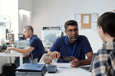 Meslektaşımla tablet tutarken teknik projeyi tartışan bir profesyonel. Arkaplan, ekip üyelerinin teknolojik aygıtların ayrıntılı planlarını ve çalışmalarını gösteriyor