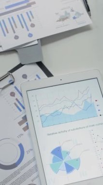 Analitik verileri gösteren dijital tabletin dikey yakın görüntüsü ve ofis masasında çizelgeleri olan yazdırılmış belgeler