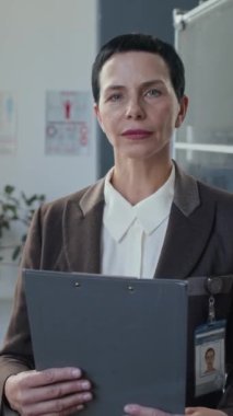 Orta yaşlı kadın tıbbi direktörün resmi. Rozetli, resmi giyinmiş, elinde not panosuyla ofise güveni tam bir şekilde bakıyor.