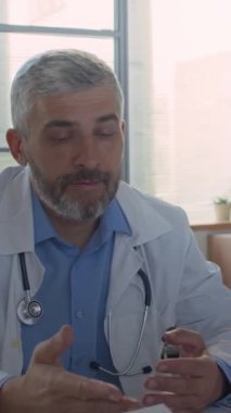 Beyaz önlüklü, gri saçlı bir erkek doktorun, ofis masasında oturup profesyonel toplantı sırasında iş arkadaşıyla yazılı belgeleri tartışması.