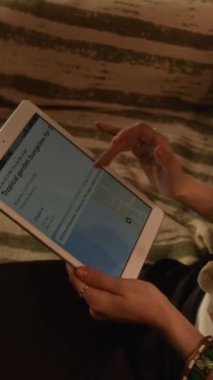 Beyaz tişörtlü, tanınmayan bir kadının plajda oturup tatil planladığı dijital tablette konaklamayı seçtiği, evdeki bungalov hakkında bilgi okuduğu dikey yüksek açı çekimi.