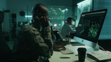 Afrika kökenli Amerikalı erkek askeri uydu operatörünün gece kumanda merkezinde çalışırken, güvenlik kameraları görüntülerini izlerken, kulaklıktaki telefona cevap verirken orta ölçekli görüntüsü.