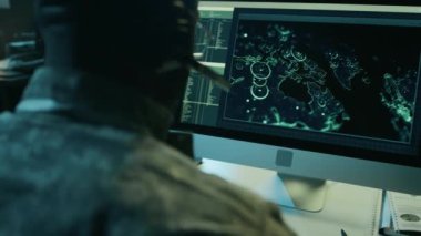 Kamuflaj üniformalı Afrikalı Amerikalı erkek askeri operatörün yakından çekilmiş arkası. Kulaklığı bilgisayarda uydu görüntülerini izliyor ve kulaklıklardan gelen çağrılara cevap veriyor.