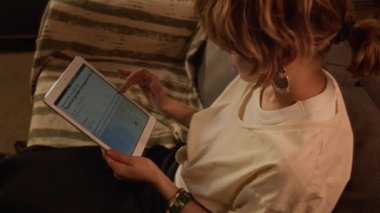 Beyaz tişörtlü, sarışın bir kadının, koltukta oturup, plajda tatil planladığı, dijital tablette konaklamayı seçtiği, evdeki bungalov hakkında bilgi okuduğu yüksek açılı bir fotoğraf.