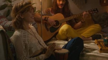 Orta boy gülümseyen beyaz adam ve kızın klasik giysiler içinde evde birlikte dinlenip gitar ve davul çalarak şarkı söylerken yatak odasındaki sisli ışıkta takıldığı bir fotoğraf.