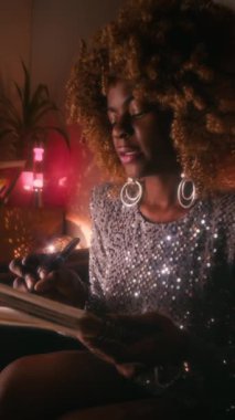 Dikey çekimde, parlak gümüş elbiseli, kabarık saçlı genç Afrikalı Amerikalı kadın şarkıcı şarkı sözleri besteleyip neon ışıklı odaya not defterine yazıyormuş.