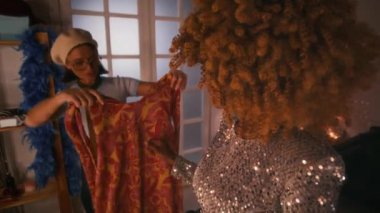 Kafkasyalı stilistin, 70 'lerin nostaljik, kıvırcık saçlı ve soyunma odasında gösteriye hazırlanan heyecanlı siyah kadın disko yıldızı için renkli bir elbise seçmesi.