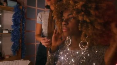 Moda tasarımcısının, parlak gümüş elbiseli ve büyük küpeli, giyinme odasında gösteriye hazırlanan Afrikalı Amerikalı kadınların kabarık saçlarını düzeltirken yakın plan fotoğrafı.