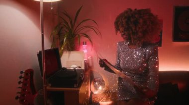 Orta ölçekli, kıvırcık saçlı, parlak gümüş elbiseli, neon ışıklı odada 70 'lerin plaklarını çalan ve heyecanlanan Afro-Amerikan bir kadın.