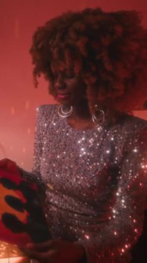 Havalı saçlı, parlak gümüş elbiseli, neon ışıklı odada 70 'lerin vinil plağını çalan modaya uygun, genç bir Afrikalı disko yıldızının dikey orta ölçekli fotoğrafı.