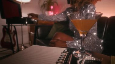 Kahve masasındaki bir bardak martiniye yakın çekim, arka plandaki koltukta oturup plakları çalan ve sanatçılar odasındaki konserden sonra dinlenen kadın ünlüler.