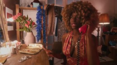 70 'lerin kıvırcık saçlı, tuvalet masasında oturan, etkin bir şekilde el kol hareketi yapan ve izlenimlerini paylaşan, konserden önce kırmızı kablolu telefonla konuşan orta sınıf siyah kadın şarkıcısı.