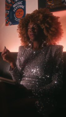 Dikey çekimde, parlak gümüş elbiseler giyen, neon ışıklı sanatçılar odasında şarkı sözü besteleyen, kabarık saçlı, genç Afrikalı Amerikalı kadın şarkıcı.