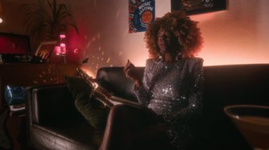 Kabarık saçlı, parlak gümüş elbiseli, deri koltukta oturan ve neon ışıklı sanatçılar odasında yeni bir şarkı besteleyen genç Afrikalı Amerikalı kadın şarkıcının tam uzun bir çekimi.