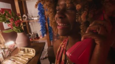 70 'lerin yakın plan çekimleri. Kabarık saçlı, tuvalet masasında oturan, gülümseyen ve izlenimlerini paylaşan genç siyahi kadın şarkıcı konser öncesi arkadaşlarıyla kırmızı telli telefonda konuşuyor.