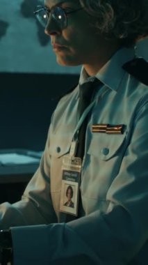 Komuta merkezinde oturan, gece vakti mavi ışıkta bilgisayar üzerinde çalışan, üniformalı ve omuz bantlı, odaklanmış beyaz kadın asker ya da polis memurunun dikey görüntüsü.