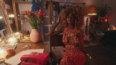 70 'lerin orta sınıf, kıvırcık saçlı, pırıl pırıl mücevherleri olan, açık hippi elbisesi içinde, martiniyle soyunma odasında oturmuş, mutlu bir şekilde gülen ve kameraya bakan siyahi kadın şarkıcının orta boy portresi.