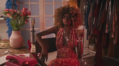 70 'lerin orta boy genç siyahi kadın portresi kabarık saçlı, ensesinde parlak mücevherler, açık hippi elbisesi içinde, tuvalet masasında bir bardak martiniyle sakince kameraya bakıyor.