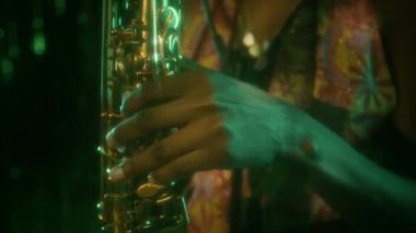 Yakın çekimde, kabarık saçlı, düğmelere basan ve saksafon çalan genç siyahi bayan müzisyenin yeşil neon ışıkta parıldayan süslemelere karşı, vücudunun üzerinde ışık huzmeleri uçuşuyor.
