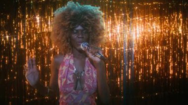 70 'lerin orta sınıf, kıvırcık saçlı, renkli elbiseli, parlak kolyeli, parıltılı arka planda mikrofona şarkı söyleyen genç siyahi kadın portresi.