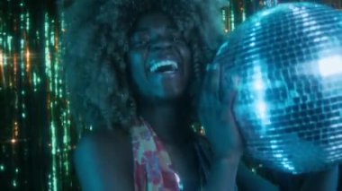 Gece kulübünde disko aynalı top tutarken parlak gülümseyen kabarık saçlı ve atmosferik 70 'li yılların nostaljik ve atmosferik kameralarına bakan Afro-Amerikan genç bayanın göğüsleri.