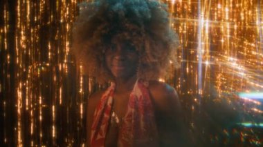 70 'li yılların Afro-Amerikan bayan disko yıldızının göğüs ölçülerinde kabarık saçlı ve açık elbiseli parlak altın renkli arka planda poz veren, gülen ve kameraya bakan, prizma