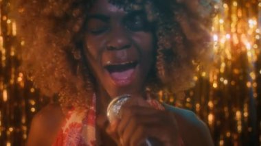 Kapalı çekimde, gece kulübünde parıltılı altın arka planda mikrofona şarkı söyleyen kabarık saçlı Afro-Amerikan genç bayanın portresi ve kameraya bakıyor.