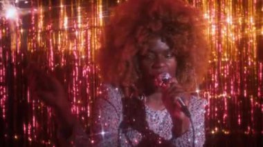 Şişkin saçlı, parlak gümüş elbiseli, mikrofona şarkı söyleyen ve parlak sahnede bengal ışığı tutan genç, şehvetli siyahi bir kadının portresi.