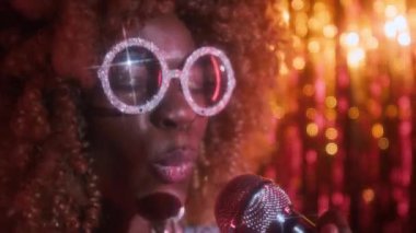 Kapalı planda, parlak gümüş gözlüklü ve elbiseli, parlak saçlı, enerjik siyahi bir bayanın 70 'lerde gece kulübünde sahnede şarkı söylerken mikrofona aktif olarak şarkı söylediği.