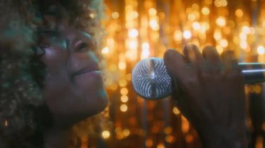 Kendine güvenen, kıvırcık saçlı, mikrofona şarkı söyleyen, 70 'li yılların nostaljik geçmişiyle eğlenen genç Afro-Amerikan bayanın yakın çekimleri.