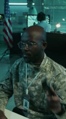 Amerikan bayrağıyla kumanda merkezinde çalışan kamuflaj üniformalı siyah erkek askerin orta boy dikey görüntüsü, sabit hattan kumandana rapor veriyor.