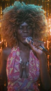 70 'li yaşlarda, kıvırcık saçlı, renkli elbiseli, parlak kolyeli, parıltılı arka planda mikrofona şarkı söyleyen, orta boylu, orta yaşlı, siyah bir kadının portresi.