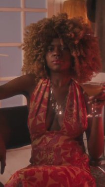 70 'lerin orta boy genç siyahi kadın portresi kabarık saçlı, ensesinde parlak mücevherler, hippi elbisesi içinde, tuvalet masasında bir bardak martiniyle sakince kameraya bakıyor.