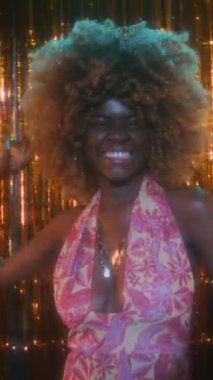 Kabarık saçlı, renkli bir hippi elbisesi giyen, 70 'li yıllarda kulüpte ışıltılı ve karizmatik bir şekilde dans eden Afro-Amerikan bir kadının dikey portresi.