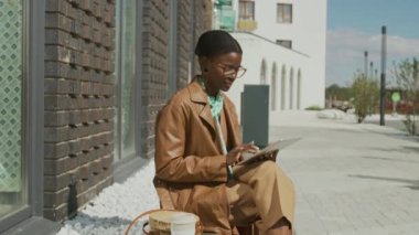 Gözlüklü, kahverengi deri ceketli ve bej pantolonlu Afro-Amerikalı genç bayan kaldırım kenarında otururken ve güneşli yaz gününde dijital tablet üzerinde çalışırken.