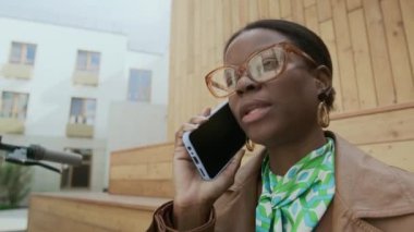 Gözlüklü ve kahverengi deri ceketli genç Afrikalı Amerikalı iş kadınının açık havada mobiletle gezerken iş arkadaşıyla telefonda konuşurken göğsü kabarmış görüntüsü.
