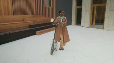 Kahverengi deri ceketli, yeşil eşarplı, iş arkadaşını bekleyen, elektrikli scooter ve bir fincan kahveyle dışarıda ofis binasının önünde duran siyahi bir iş kadını.