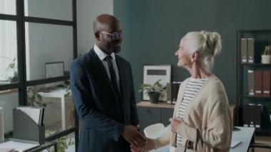 Siyahi finansal danışmanın portresi resmi giyimde yaşlı beyaz bir kadınla el sıkışıyor, kameraya poz veriyor ve ofisteki muayeneden sonra gülümsüyor.