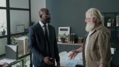 Beyaz saçlı ve sakallı yaşlı beyaz bir adamın portresi Afrika kökenli Amerikalı bir finans danışmanıyla el sıkışıyor ve ofiste başarılı bir muayeneden sonra kameraya gülümsüyor.