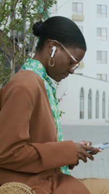 Gözlüklü ve kahverengi ceketli Afro-Amerikalı iş kadınının dikey yan görüntüsü. Kulaklıkları sokakta bankta oturuyor ve yaz günü telefonda yeni bir projeye bakıyor.
