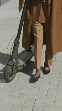 Siyah iş kadını, gözlüklü, kahverengi ceketli ve yeşil başörtüsü takmış sokakta yürürken, yanında elektrikli scooter iterken, iş ortağıyla konuşurken,