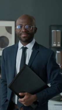 Resmi giyinmiş Afro-Amerikan iş adamının dikey portresi. Ofiste duruyor, klasörü tutuyor ve gülümseyerek kameraya bakıyor.